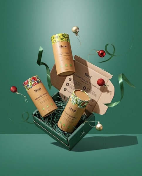 A studio-shot, 4:5 ratio creative featuring three cylindrical superfood supplement containers floating inside an open product box, accented by holiday ornaments and ribbon.